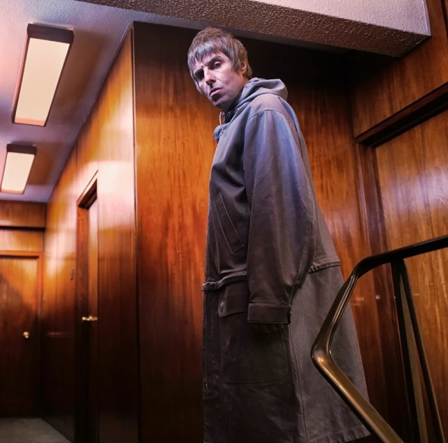Adult male in long coat walks down wood-paneled hallway with overhead lighting, captured in low-angle medium shot.