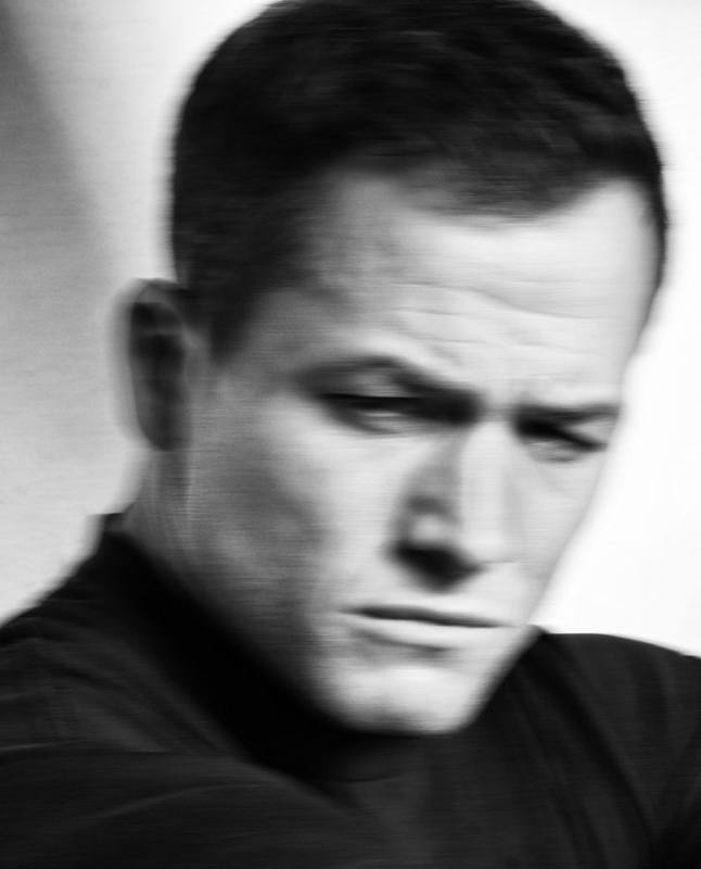 Close-up portrait of actor Daniel Craig with a serious expression, wearing a black suit and bow tie, against a dark background.