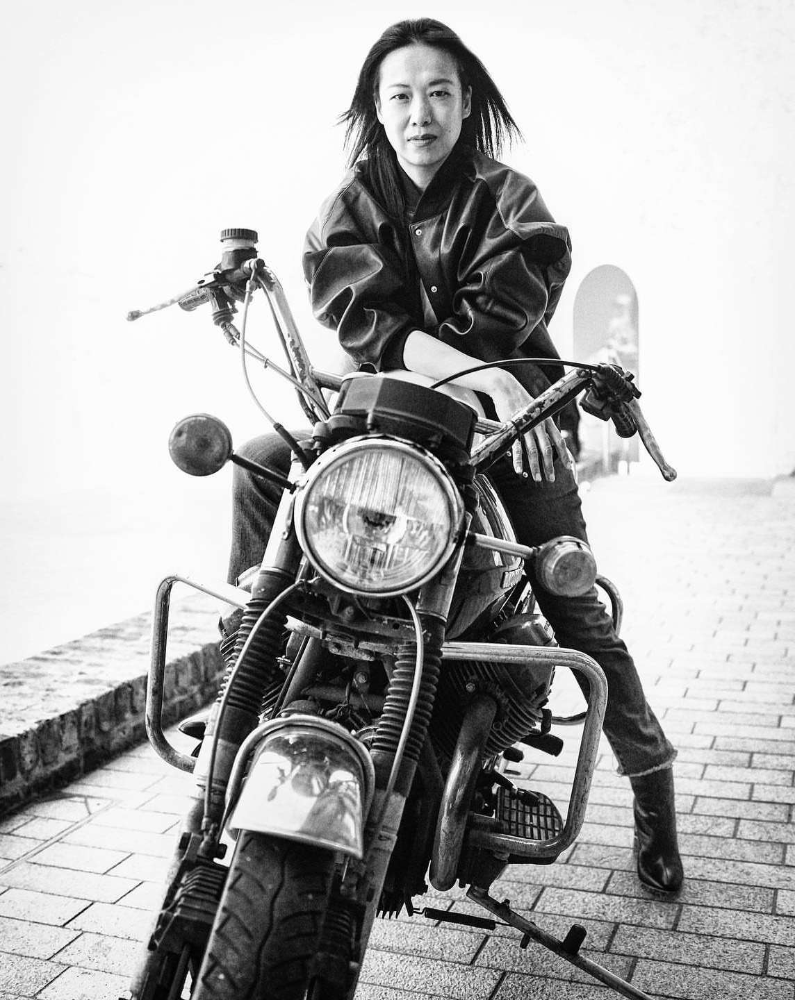 Monochrome portrait of an adult woman seated on a motorcycle, wearing a jacket, jeans and boots on a brick-paved street. 