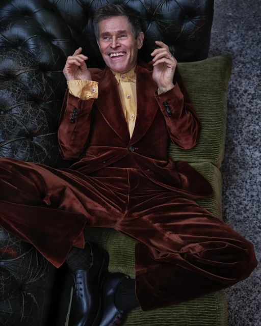 Portrait of an adult male lying on a couch wearing a brown velvet suit and black shoes, smiling with his hands raised up.