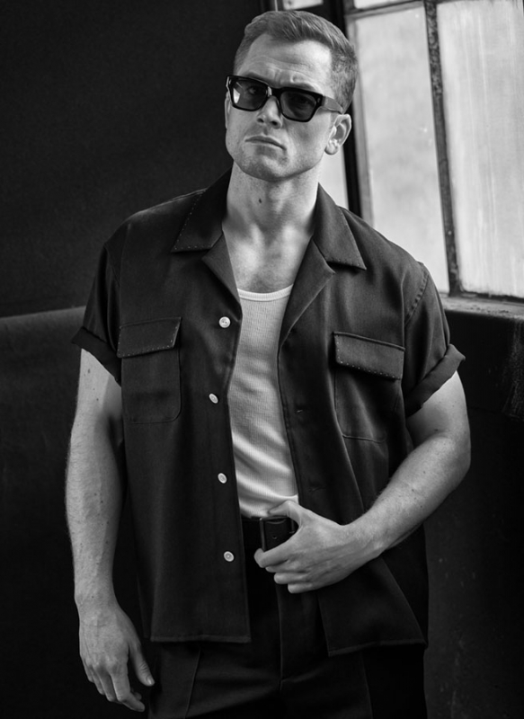 Monochrome portrait of a man wearing sunglasses and an open camp shirt, leaning against a wall with a serious expression.