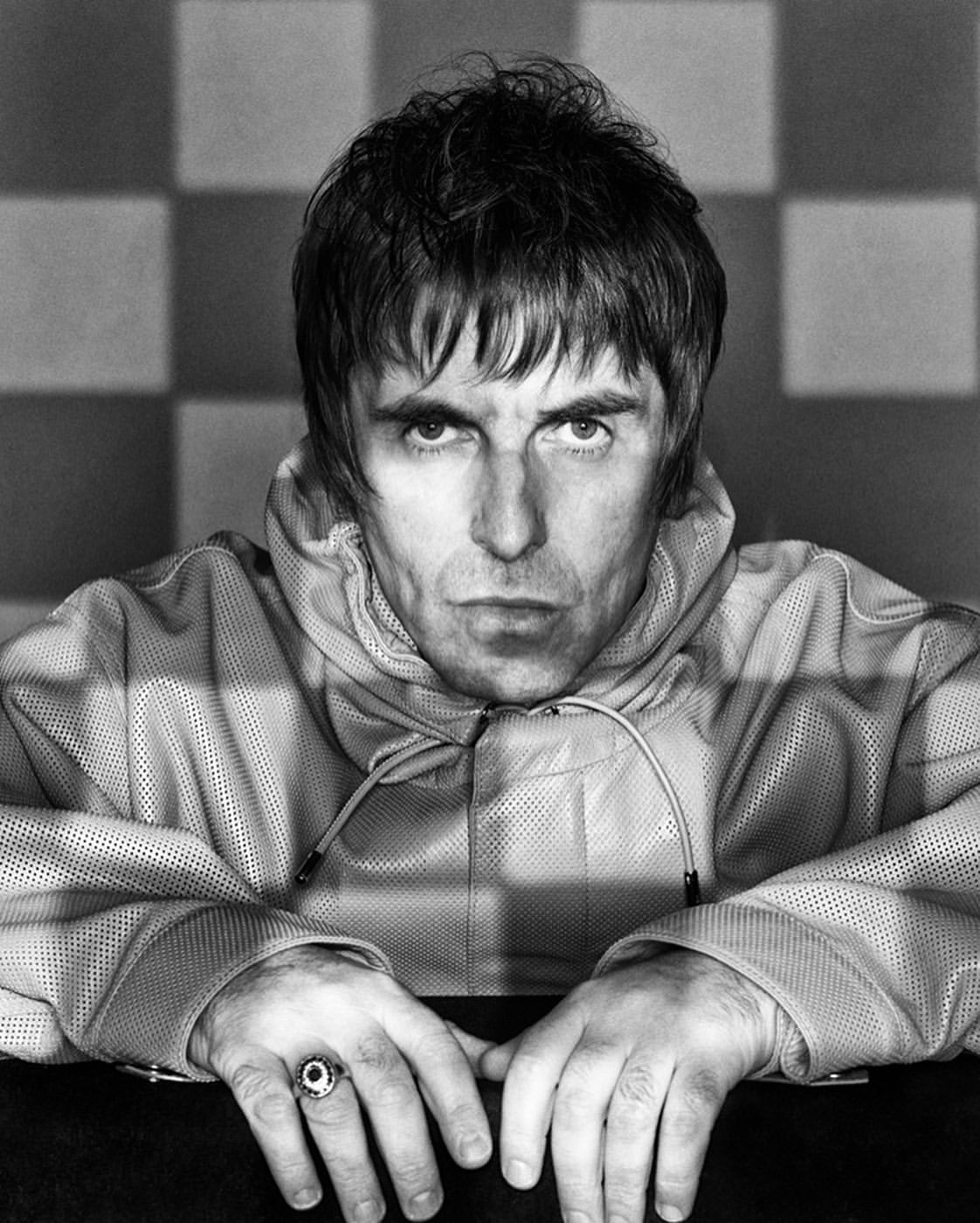 Portrait of Liam Gallagher in a zipped hooded top leaning forward with a ring on his finger against a checkered background