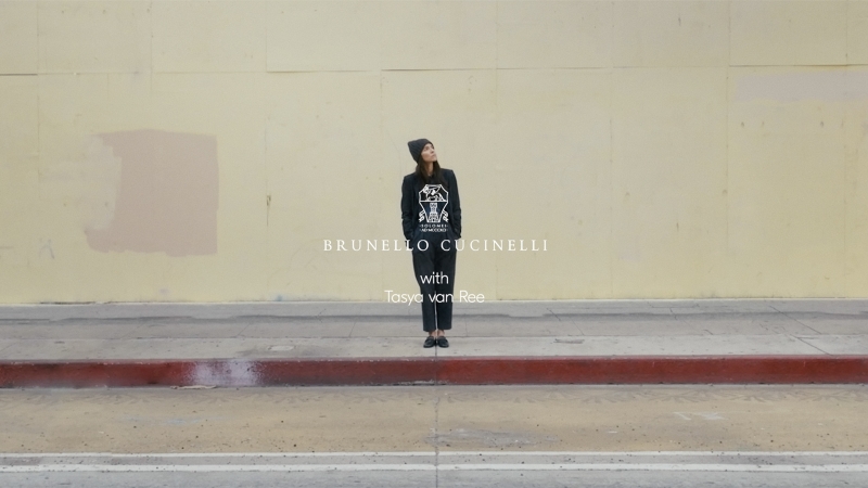 Woman in dark streetwear stands on sidewalk in front of aged beige wall, looking up; fashion shot with brand logo text overlay.