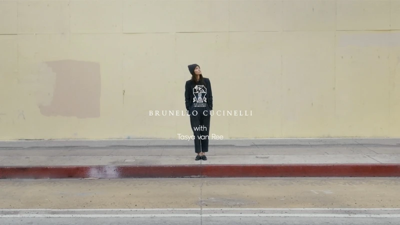 Woman in dark streetwear stands on sidewalk in front of aged beige wall, looking up; fashion shot with brand logo text overlay.