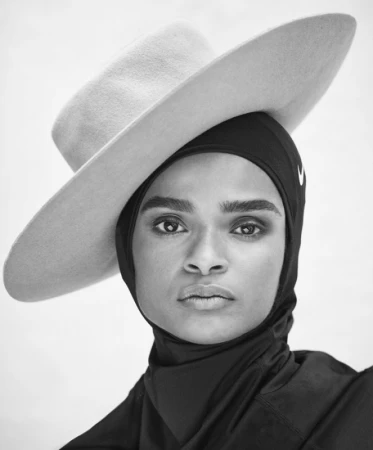 Close-up portrait of a woman wearing a black athletic hijab and a wide-brimmed hat against a light background.
