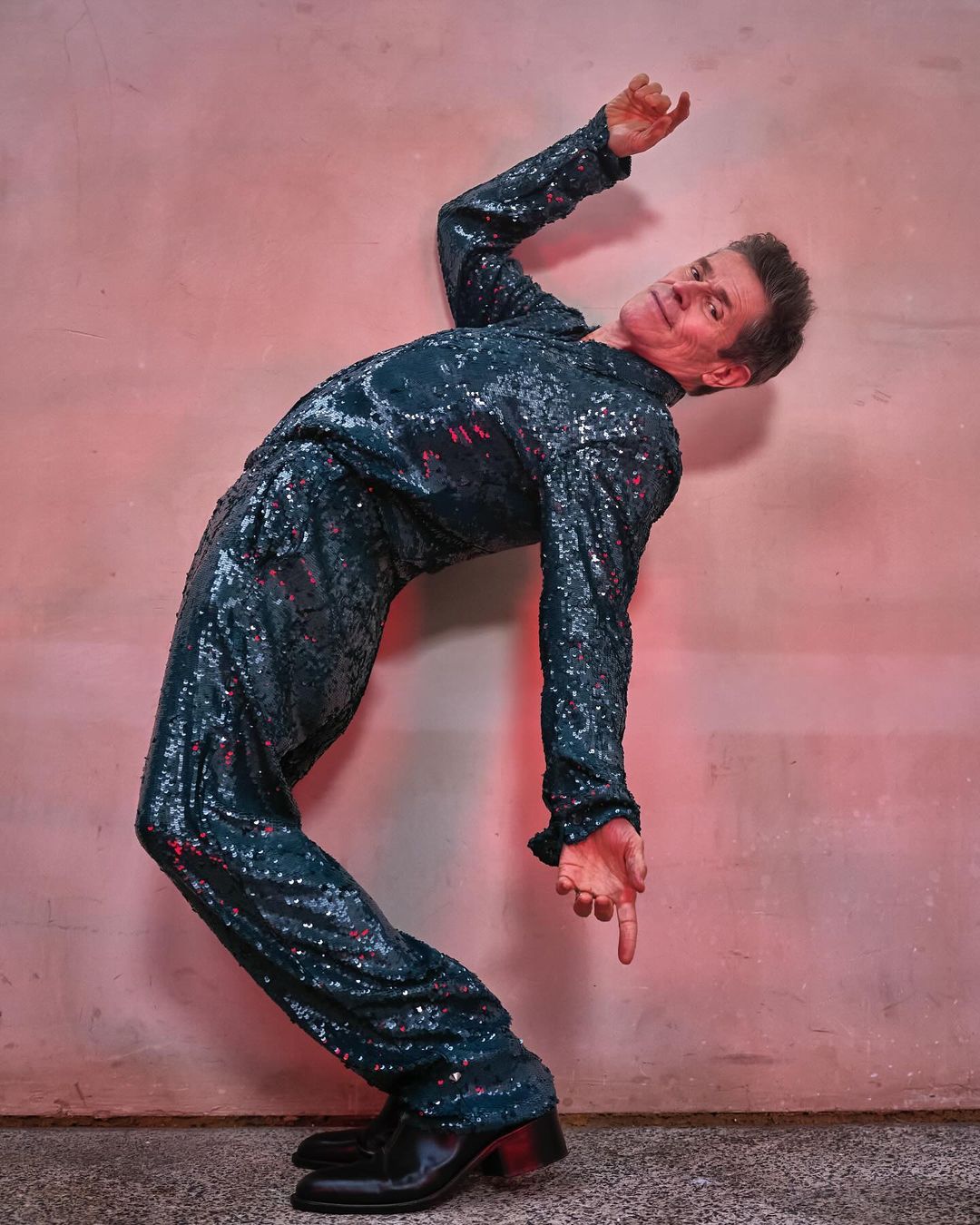 Man in a sparkling teal suit posing and pointing down against a light pink wall with neutral grey flooring in an indoor studio setting.