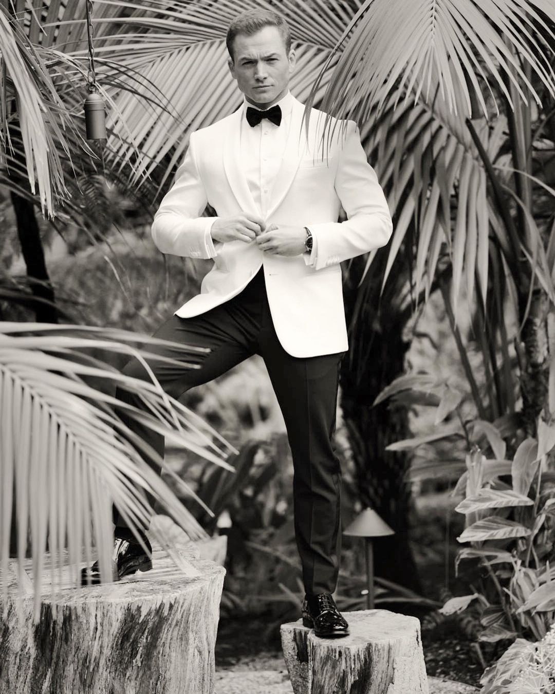 Black and white portrait of a man in a white tuxedo and bow tie standing in a lush, tropical environment with tall palm fronds.