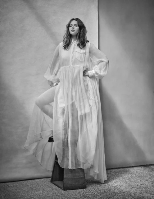 Monochrome portrait of a woman in a sheer white dress posing in a photo studio, with soft lighting and a textured backdrop.