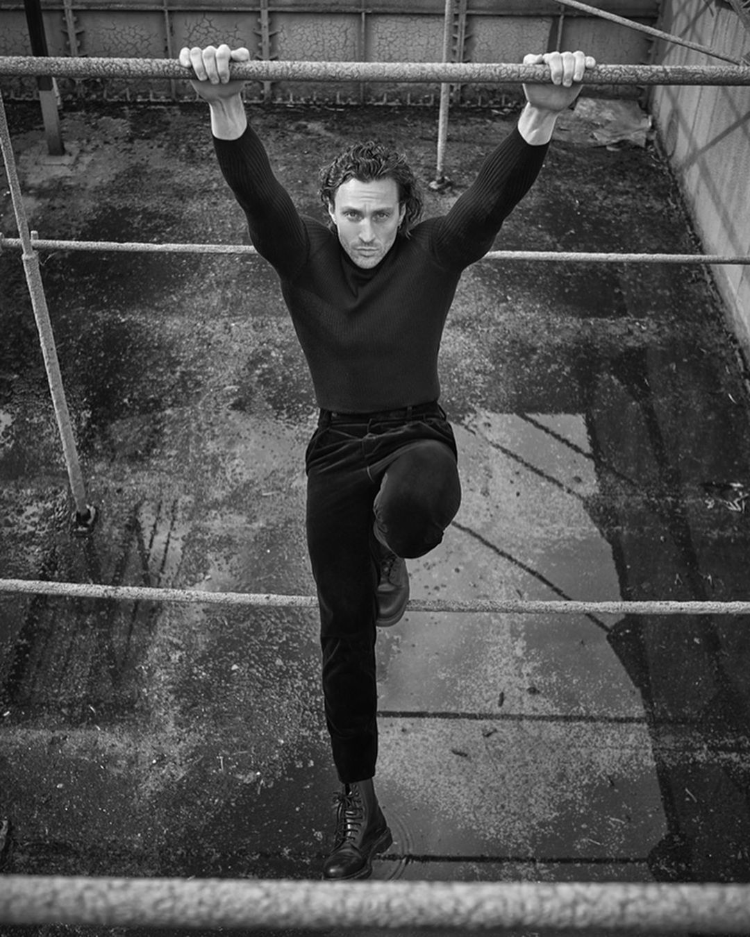 Black and white portrait of a man hanging from metal bars, arms extended overhead, one leg extended forward, wearing dark clothing.