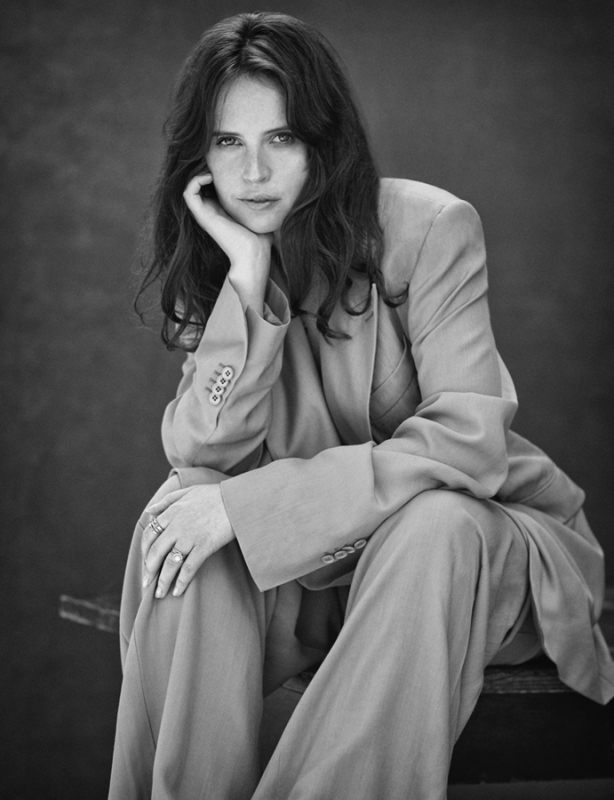 Monochrome portrait of a woman in a suit sitting with her head resting on her hand, looking at the camera against a dark background.