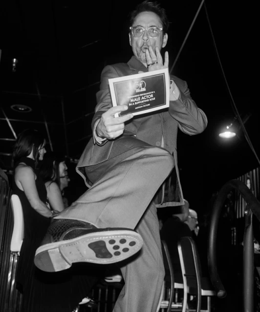 Man in suit holds award and kicks leg out. Black and white image features celebration, formal wear, and stage presence.