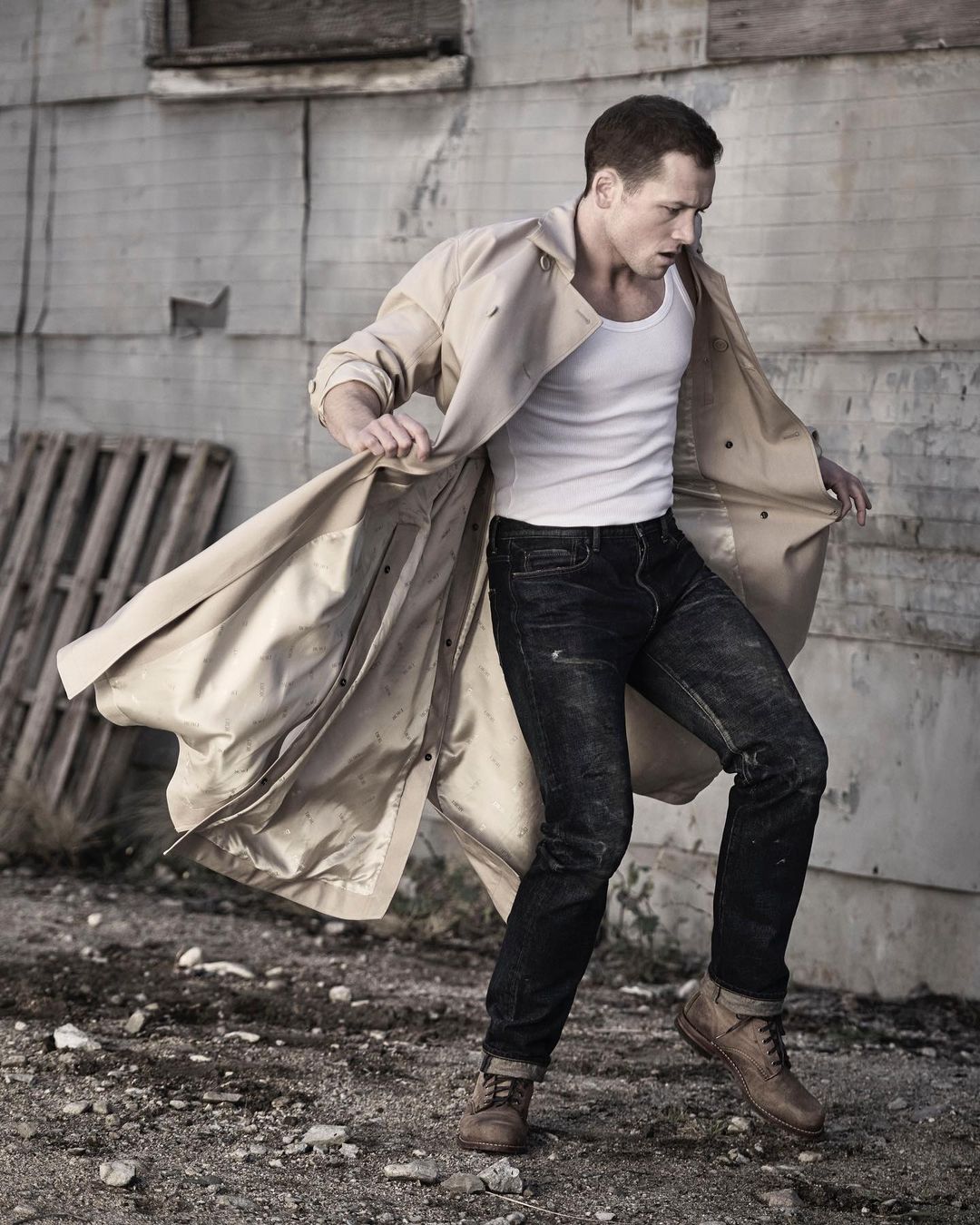 Adult male poses mid-motion in an urban alleyway, wearing a light trench coat, white t-shirt, jeans, and brown work boots in outdoor setting.