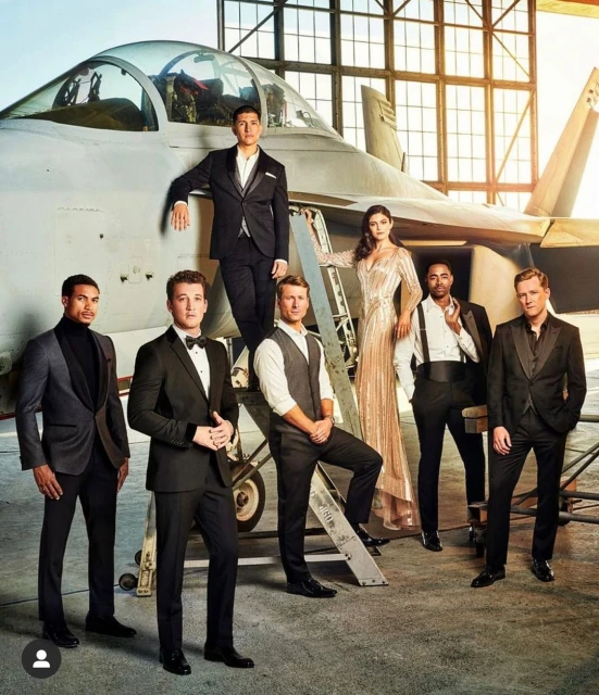 Group of six people in formal wear posing in front of a military jet; the woman wears a long dress and the men wear suits.