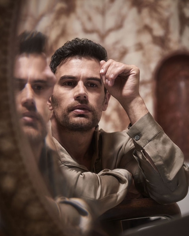 Portrait of a brunette adult male in a collared shirt, looking at the camera, reflected in a mirror with a patterned backdrop.