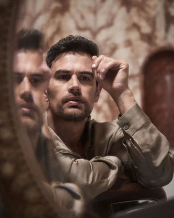 Portrait of a brunette adult male in a collared shirt, looking at the camera, reflected in a mirror with a patterned backdrop.