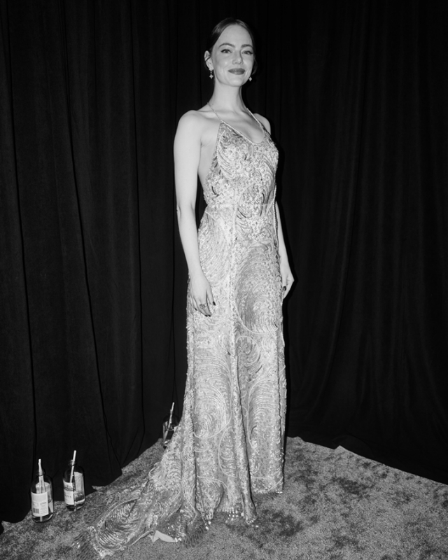 Emma Stone poses in an elegant beaded gown against a black backdrop, showcasing a full-length portrait in monochrome. Soft lighting enhances the gown's intricate details.