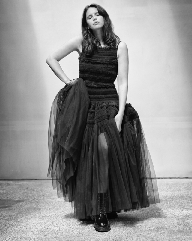 Monochrome portrait of a woman in a black dress and boots. She is posing indoors with a neutral expression and soft lighting.
