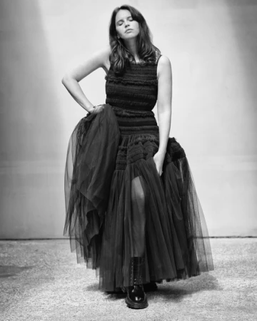 Monochrome portrait of a woman in a black dress and boots. She is posing indoors with a neutral expression and soft lighting.
