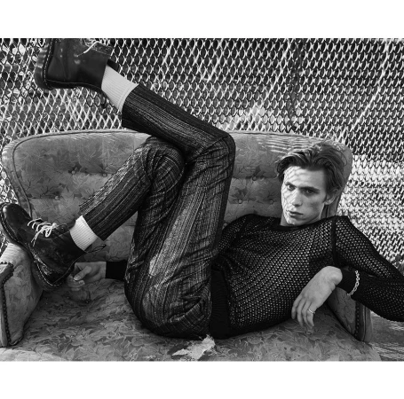 Monochrome portrait of a young man reclining on a patterned vintage sofa with his legs kicked up wearing laced boots.