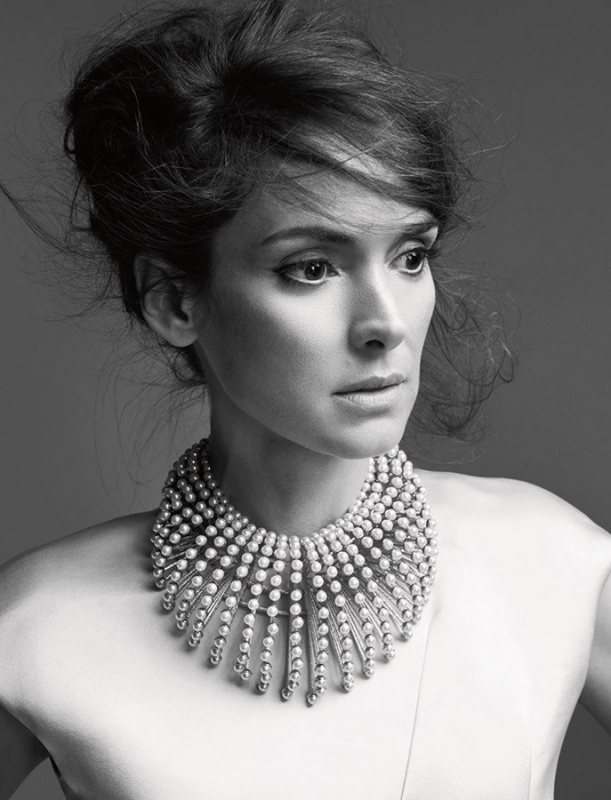 Black and white portrait of Winona Ryder wearing pearl necklace against solid grey background. Hair is styled up and away from face.