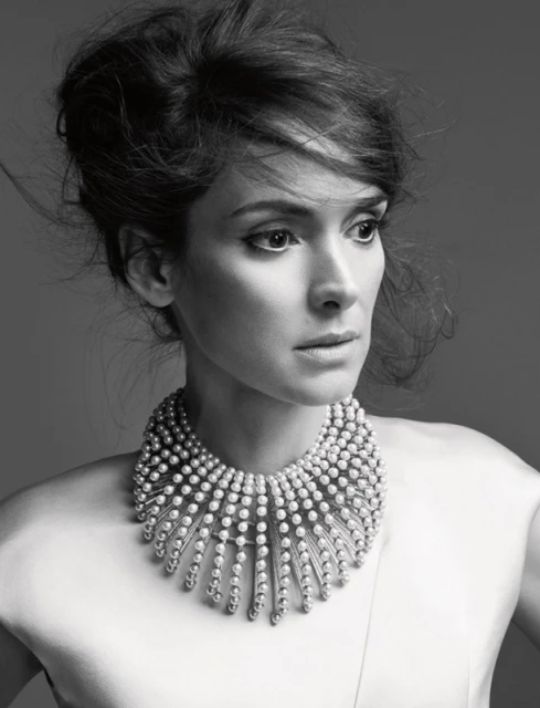 Black and white portrait of Winona Ryder wearing pearl necklace against solid grey background. Hair is styled up and away from face.