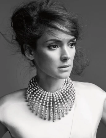 Black and white portrait of Winona Ryder wearing pearl necklace against solid grey background. Hair is styled up and away from face.