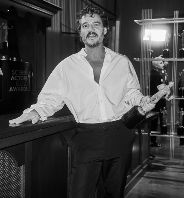 Black and white shot of a man in an unbuttoned white shirt and dark pants holding an award, leaning against a counter in a room.