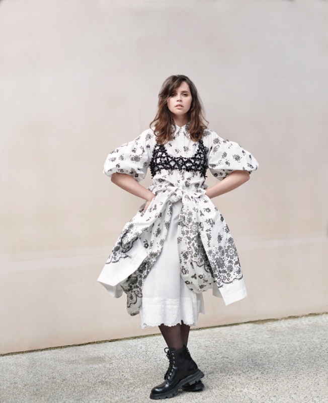 A woman in a white floral dress with black combat boots stands in front of a light tan wall, looking at the camera.