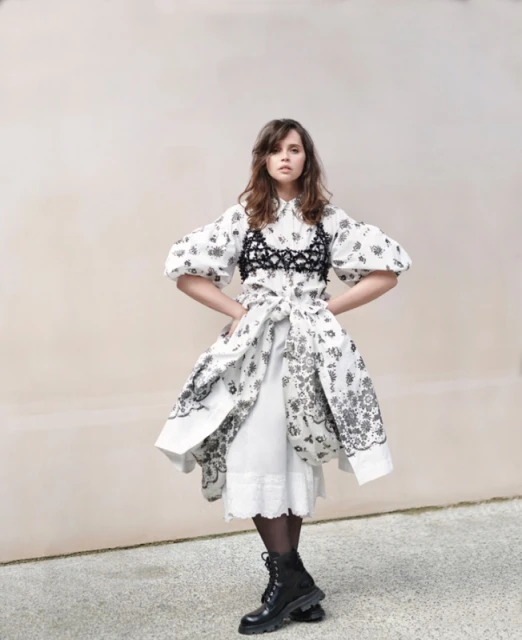 A woman in a white floral dress with black combat boots stands in front of a light tan wall, looking at the camera.