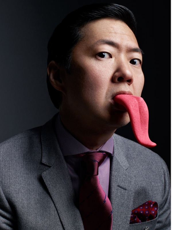 Portrait of an Asian adult male wearing a grey suit with a large pink fake tongue sticking out of his mouth against a dark backdrop.
