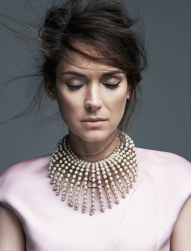 Portrait of Winona Ryder with eyes closed, wearing a layered pearl collar necklace and pink top against a grey backdrop.