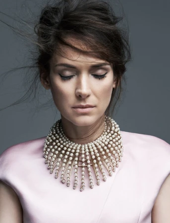 Portrait of Winona Ryder with eyes closed, wearing a layered pearl collar necklace and pink top against a grey backdrop.