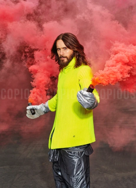 Portrait of Jared Leto holding two red smoke flares against a red smoky background, wearing a neon yellow sweater and silver pants.