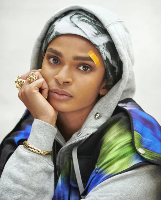 Portrait of a woman wearing a hoodie and jewelry with a bandage on her forehead against an off-white background.