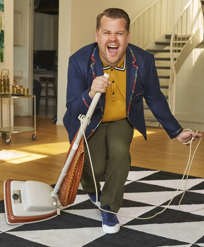 James Corden sings into a vintage vacuum cleaner on a black and white geometric patterned rug in a bright room.