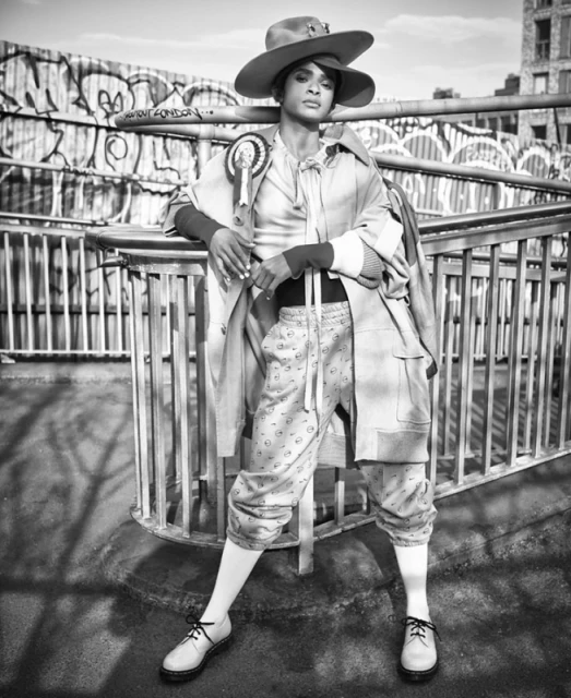 A black and white portrait of a woman in a hat and overcoat posing on a bridge with graffiti in the background and wearing formal shoes.