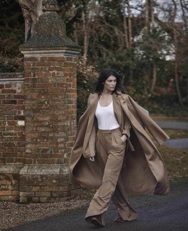 Adult woman walking in a fashionable tan pantsuit and white top outdoors near a brick gate with an overcast sky in the background.