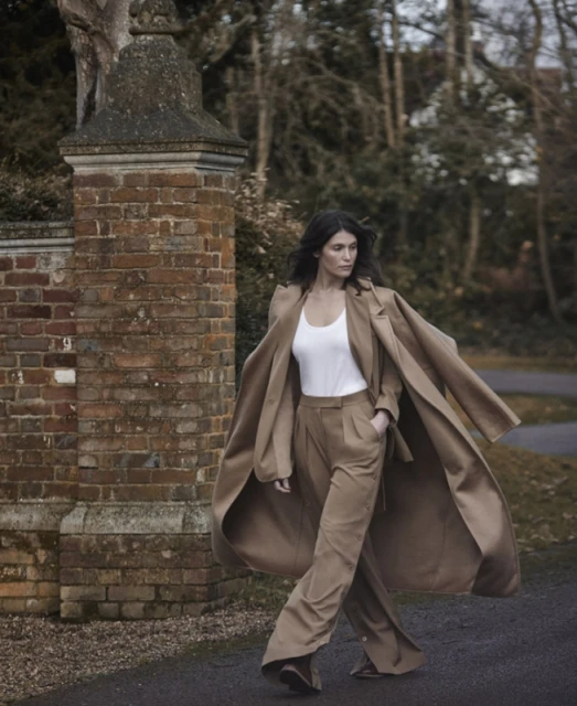 Adult woman walking in a fashionable tan pantsuit and white top outdoors near a brick gate with an overcast sky in the background.