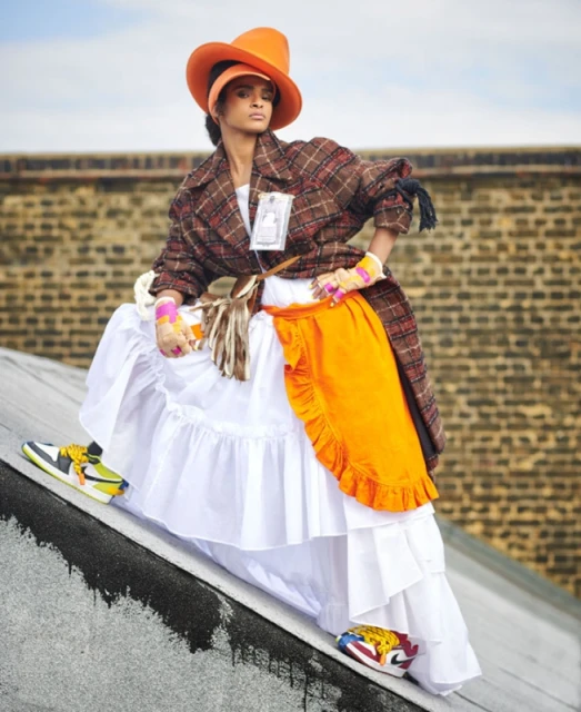 A stylish woman in a hat, coat, and sneakers poses on a rooftop, showcasing colorful, avant-garde fashion with an urban background.
