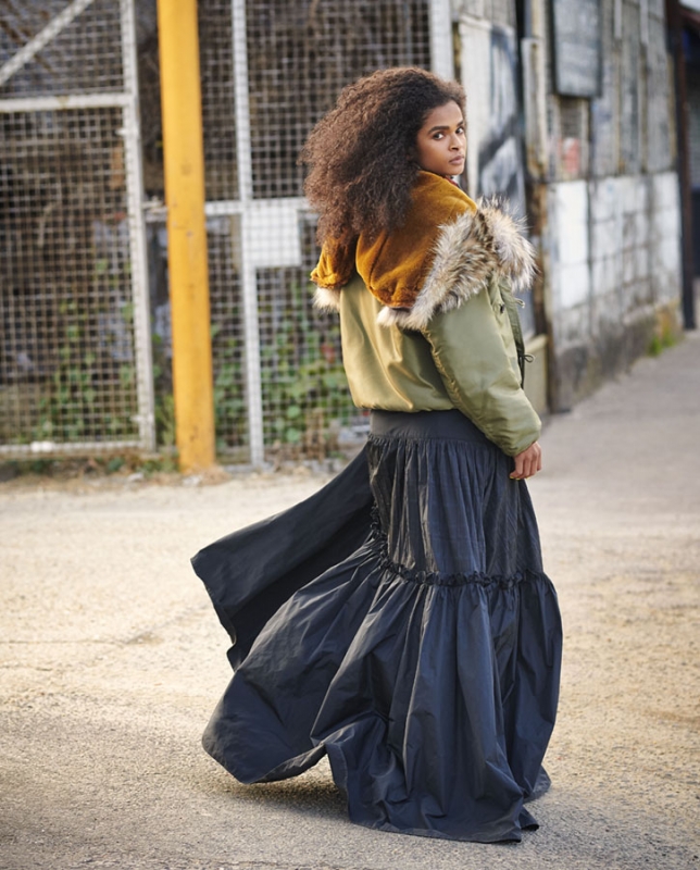 Woman with curly hair posing in a puffy olive jacket with fur hood, and a long black skirt on an urban street with metal fence.