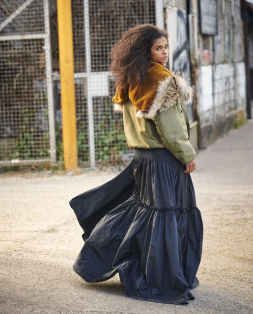 Woman with curly hair posing in a puffy olive jacket with fur hood, and a long black skirt on an urban street with metal fence.