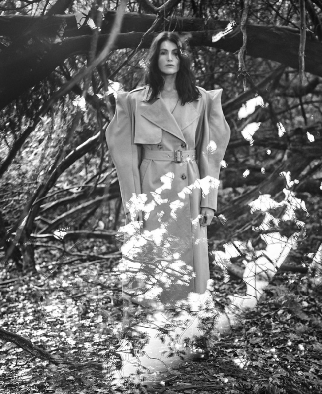 A woman in a belted trench coat stands amidst a monochromatic forest setting with scattered leaves and visible branches.