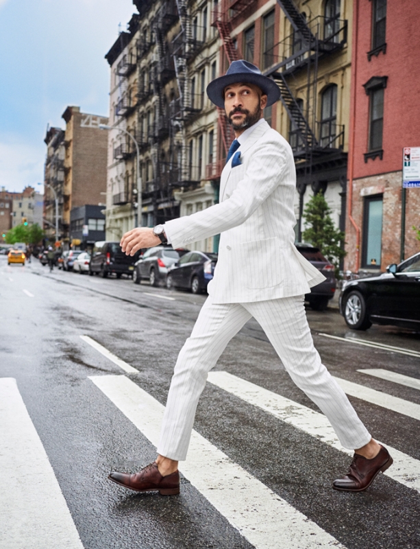 Man in a white suit walks across a zebra striped crosswalk in New York City with parked cars and buildings in the background.