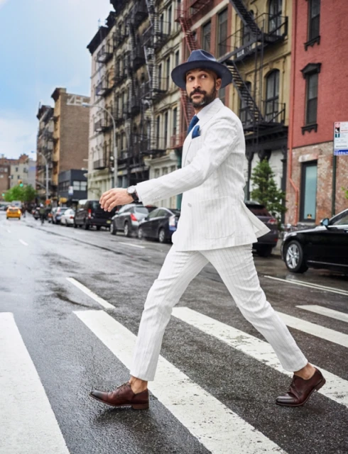 Man in a white suit walks across a zebra striped crosswalk in New York City with parked cars and buildings in the background.
