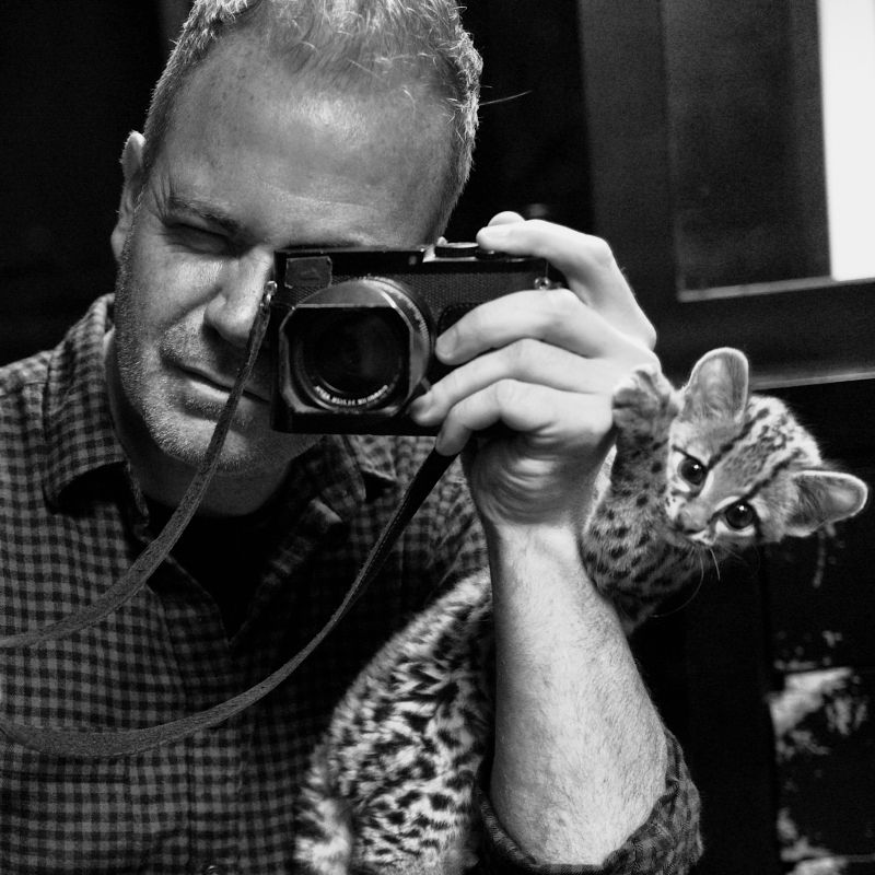 Black and white portrait of a man holding a camera with a cat perched on his shoulder looking at the camera.
