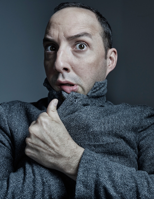 Portrait of a fair-skinned adult male in a grey jacket with a surprised facial expression against a dark grey background indoors.