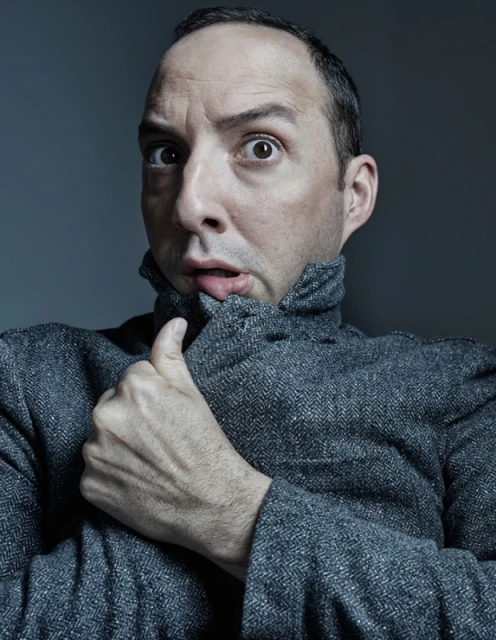 Portrait of a fair-skinned adult male in a grey jacket with a surprised facial expression against a dark grey background indoors.