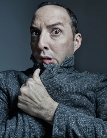 Portrait of a fair-skinned adult male in a grey jacket with a surprised facial expression against a dark grey background indoors.