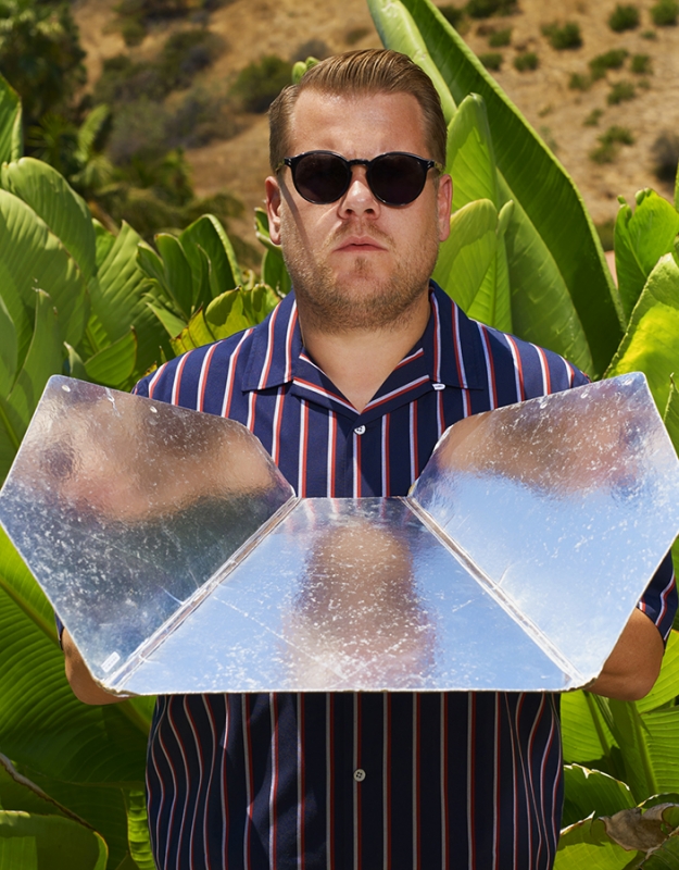 An adult male wearing sunglasses and striped shirt holding a silver, foldable solar cooker outdoors in front of green foliage.