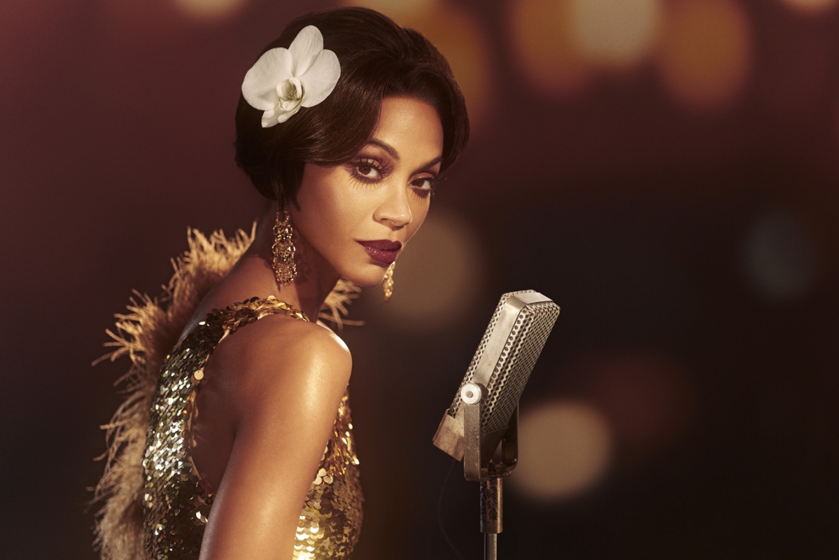 Close-up portrait of Zoe Saldana with dark hair, white orchid hairpin, and sequined gold dress singing into a vintage microphone.